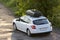 New clean white modern car with black roof luggage box container moving along empty asphalt road in bad condition by green trees a