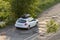 New clean white modern car with black roof luggage box container moving along empty asphalt road in bad condition by green trees