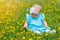 A newborn baby girl in a blue dress sits on a field with yellow dandelions. Baby 8-10 months old plays with soap bubbles