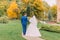 Newly married couple strolling in park. Groom is gently holding hand of his elegant bride