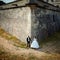 Newlyweds are holding hands while standing in the corner of the huge walls of the old baroque castle.