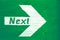 `Next` text written on a white directional arrow painted on a green wooden signboard background.