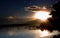 Next to the pier, the sun over the mountains, a sundog and clouds are reflected in the calm lake