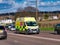 An NHS ambulance travelling at speed on a road in a semi-urban area of England, UK.