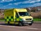 An NHS ambulance travelling at speed on a road in a semi-urban area of England, UK.