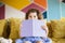 Nice baby girl with dark curly hair in blue dress happily looking in camera while covering mouth with book on sofa at