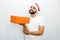 Nice bearded man in red hat stands and holds orange box with present in one hand. He looks on camera and smiles a bit