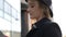 Nice blond girl in black coat and hat listens to music with smartphone headphones against city street slow motion