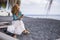 Nice caucasian lady sitting on a seat at the black vulcanic beach looking at the ocean and waves while check the smartphone with