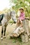 A nice little girl with light curly hair in a vintage plaid dress and a straw hat and a gray horse. Horses a