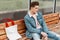 Nice stylish young man in trendy blue youth casual jeans clothes sitting on wooden bench and waiting for public transport.