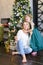 Nice woman with bengal light wearing jeans and sitting with daughter near Christmas tree.