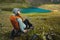 nice woman in a panama with a backpack sits on a stone, having a rest after trekking in the mountains, near the lake