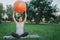 Nice young pregnant woman sit in lotus pose in park. She hold big orange fitness ball with hands and look to right. She