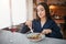 Nice young woman sit at table and eat salad. She hold both fork and knife. Model look down. Glass of red wine stand on