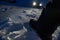 At night. Detail of leg and boot stepping on the snow with many footprints around and an all-terrain car with the lights on,