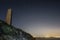 Night image of a beach with a stone monolith, lights and city in the background and a line of a star forming a circumpolar, in