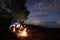 Night summer camping on shore. Group of young tourists around campfire near tent under evening sky