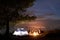 Night summer camping on shore. Group of young tourists around campfire near tent under evening sky