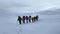 Nine hikers with large backpacks are walking along a snow-covered mountain trail.