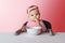 Nine-month-old baby girl in pink bandage sits at white table in highchair, eats herself with spoon from bowl. Pink background