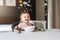 Nine-month-old smiling baby girl sits at white table in highchair and eats herself with spoon from bowl. Blurred background