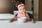 Nine-month-old smiling baby girl sits at white table in highchair and eats herself with spoon from bowl. Blurred background