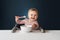 A nine-month-old smiling baby girl sits at a white table in a highchair and eats herself with a spoon from a bowl. Dark background