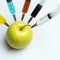 Nitrates, pesticides, fungicides and other chemicals are injected into a green apple with a syringe. Nearby is a