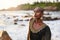 Non-binary black person in luxury dress sits on rocks in ocean. Trans ethnic fashion model wearing jewelry in posh gown