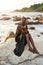 Non-binary black person in luxury dress sits on rocks in ocean. Trans ethnic fashion model wearing jewelry in posh gown
