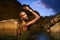 Non-binary person of color poses in water, shows jewelry inside scenic creek at night. Lgbtq ethnic graceful fashion