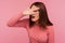 Nosy brunette woman in pink sweater spying through hole in fingers pretending to close eyes with hand, hiding to watch secrets