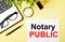 NOTARY PUBLIC is written in red on a white piece of paper on a light yellow background next to a laptop, pen, magnifying glass,