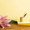 Notebook, colored pencils, flower on wooden desk. Yellow background