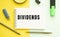 Notebook with text DIVIDENDS on office table with office supplies. Yellow color background