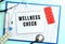 A notepad with the text WELLNESS CHECK lies on a medical clipboard with a stethoscope and pills on a blue background.