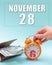 November 28th. Hand holding an orange alarm clock, a wallet with cash and a calendar date. Day 28 of month.