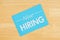 Now hiring on a blue card with a pen on textured wood background