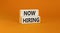 Now hiring symbol. Concept words Now hiring on beautiful wooden block. Beautiful orange table orange background. Business