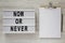 `Now or never` word on lightbox, clipboard with sheet of paper on a white wooden surface, top view. Space for text