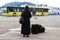 Nun traveling with suitcases and luggage. A woman in black monastic clothes is waiting for a bus on the waterfront of