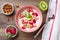 Oatmeal porridge with slices of kiwi, strawberries, almonds in pink bowl, spoon, napkin with red stripes on wooden background