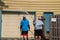 Obese Caucasian man is spray pressure washing the siding on his house. His son is holding the hose and watching