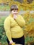 Obese smiling girl in yellow clothes stands on background of autumn trees and looks away