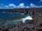 Ocean waves breaking on the rocky coast of hardened lava with caverns and cavities. Deep blue sky with white clouds and mountains