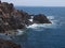 Ocean waves breaking on the rocky coast of hardened lava with caverns and cavities. Mountains and volcanoes on the horizon