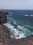 Ocean waves breaking on the rocky coast of hardened lava with caverns and cavities. Mountains and volcanoes on the horizon