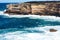 Oceanside rocky sandstone cliff with blue sea water waves creating whitewash against coastline and clear sky in background