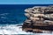 Oceanside rocky sandstone cliff with blue sea water waves creating whitewash against coastline and clear sky in background